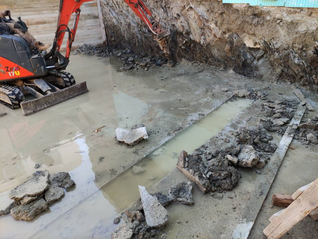 A small excavator digging a trench at the bottom of the pit, in the water.