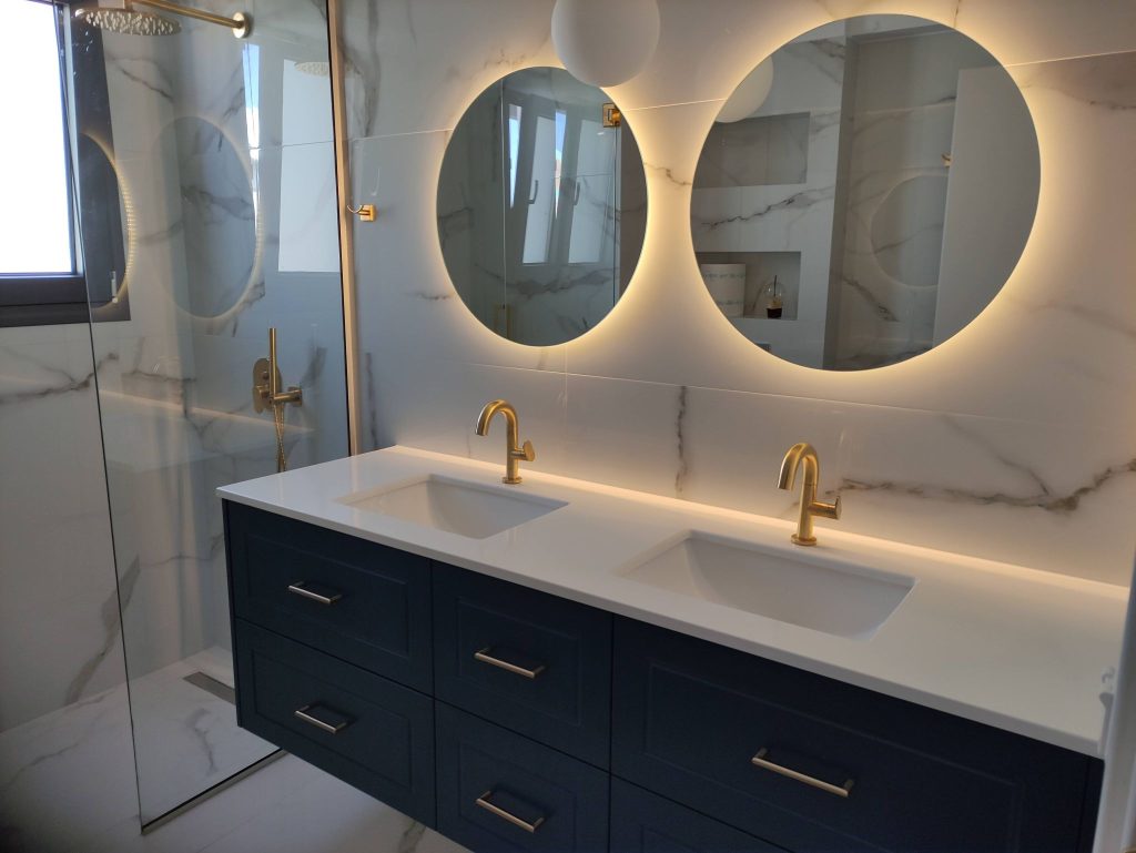A bathroom with a double basin, round illuminated mirrors and gold taps.