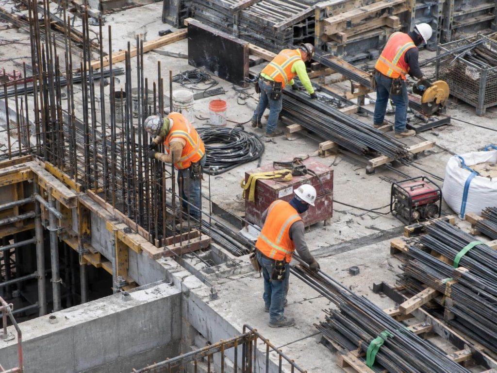 A crew in helmets and hi-vis vests working on column and slab reinforcement on a building site, with bundles of reinforcing bars around them.